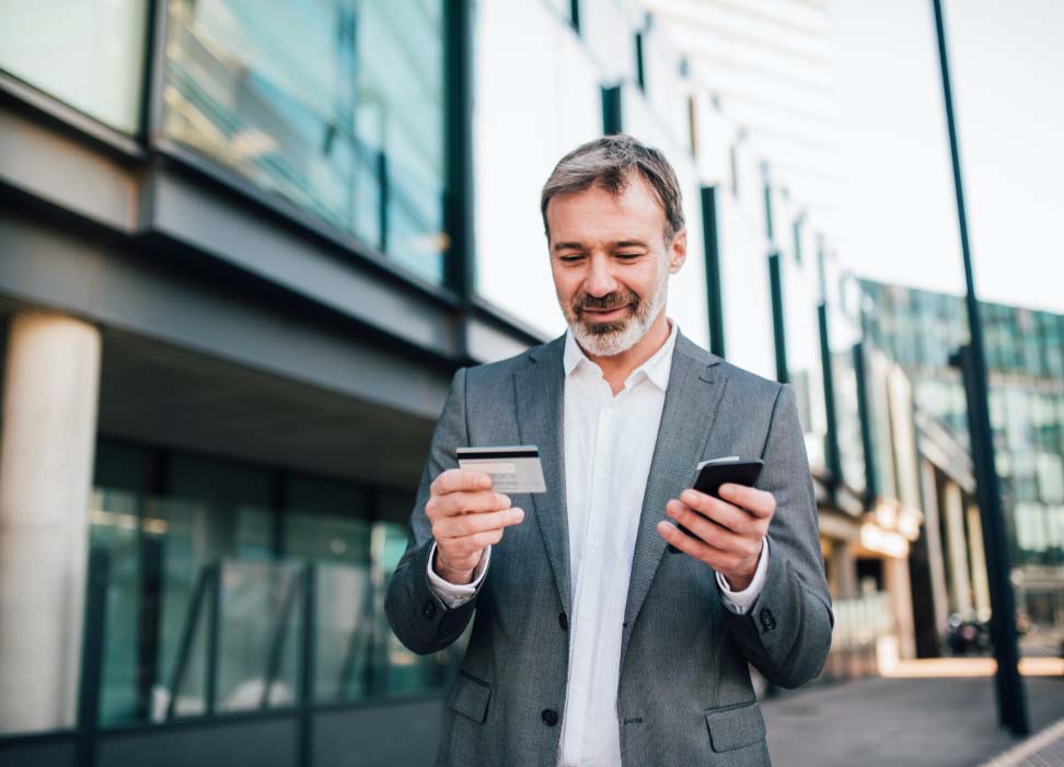 Man standing in street holding mobile device while looking at credit card.